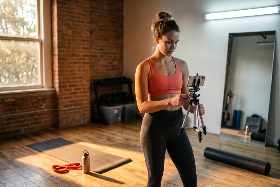 Fitness coach filming a workout tutorial in a bright, clean home gym setup with a ring light and phone tripod